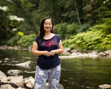 Headshot of Rae Tajiri. She is standing next to a river and trees.